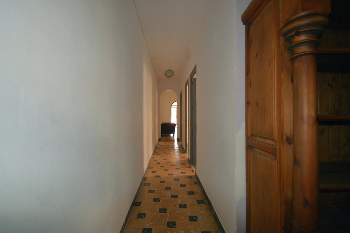 Long couloir avec un sol en carrelage à motifs et des murs blancs, menant à une ouverture en arche et un meuble en bois massif aux détails sculptés.