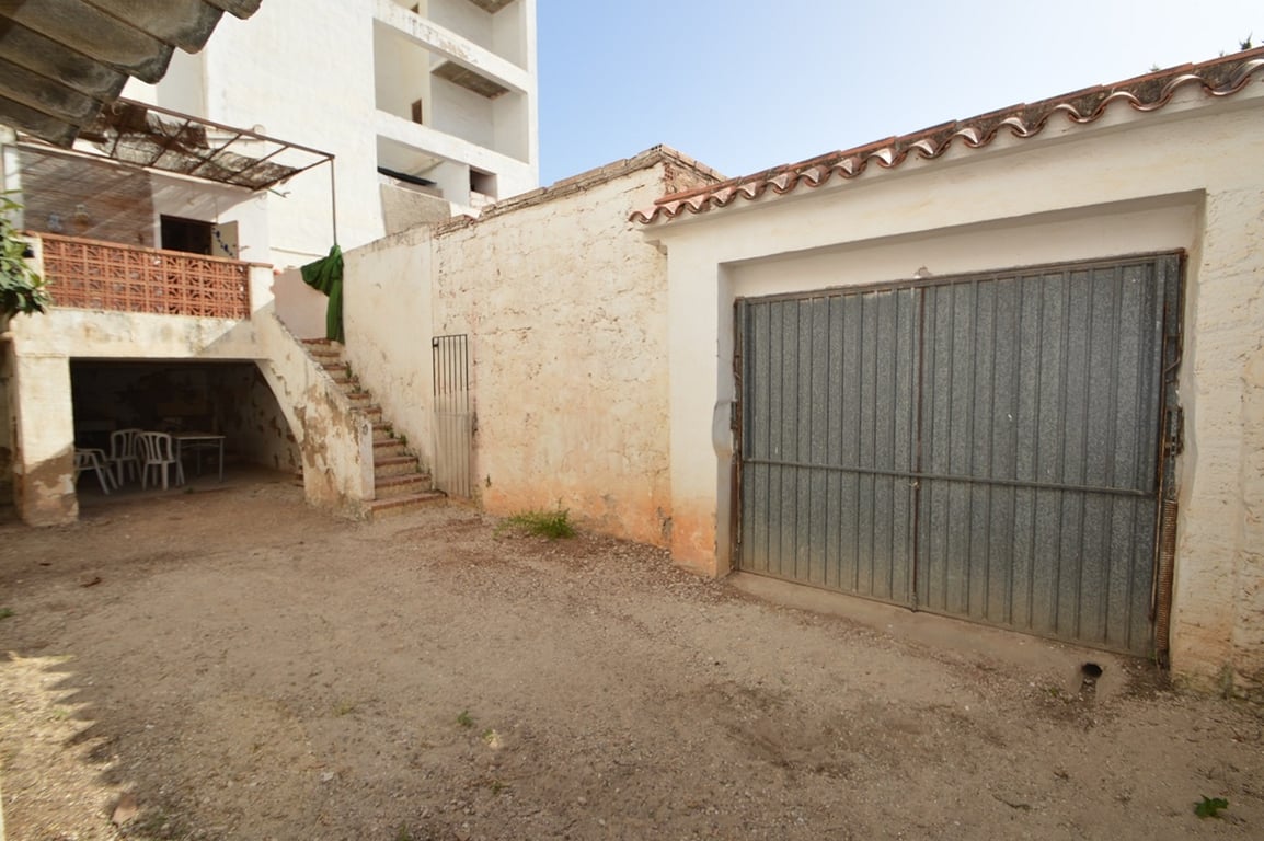 Patio arrière avec porte de garage métallique pour parking privé, escalier extérieur menant à la terrasse supérieure et espace couvert polyvalent.