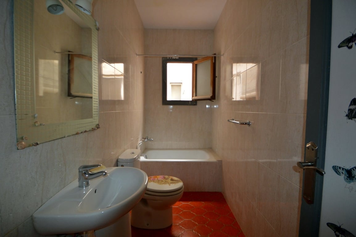 Salle de bain équipée d'un sol en tomettes hexagonales rouges, de murs carrelés beiges, d'un lavabo sur colonne et d'une baignoire encastrée avec fenêtre de ventilation.