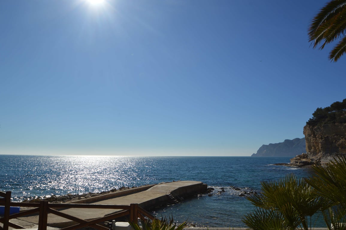 Vue panoramique sur la mer Méditerranée depuis la côte de Benissa, avec une jetée en béton et des falaises escarpées sous un soleil radieux.