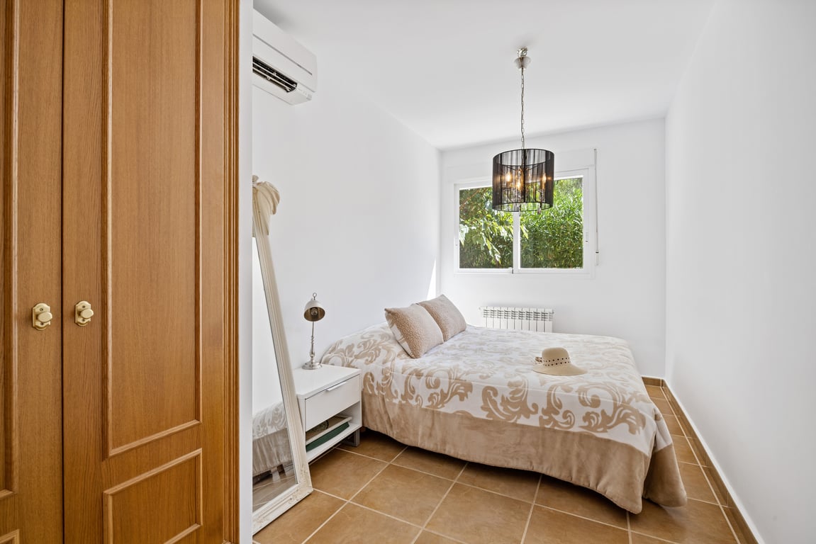 Chambre lumineuse avec placards intégrés en bois, climatisation réversible et sol en grès. La fenêtre donne sur la verdure et dispose d'un radiateur attenant.