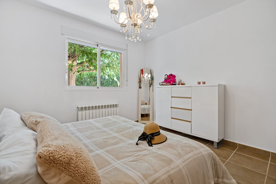 Chambre lumineuse avec sol en grès, fenêtre à double vitrage vue jardin, radiateur de chauffage central et lustre classique à pampilles.
