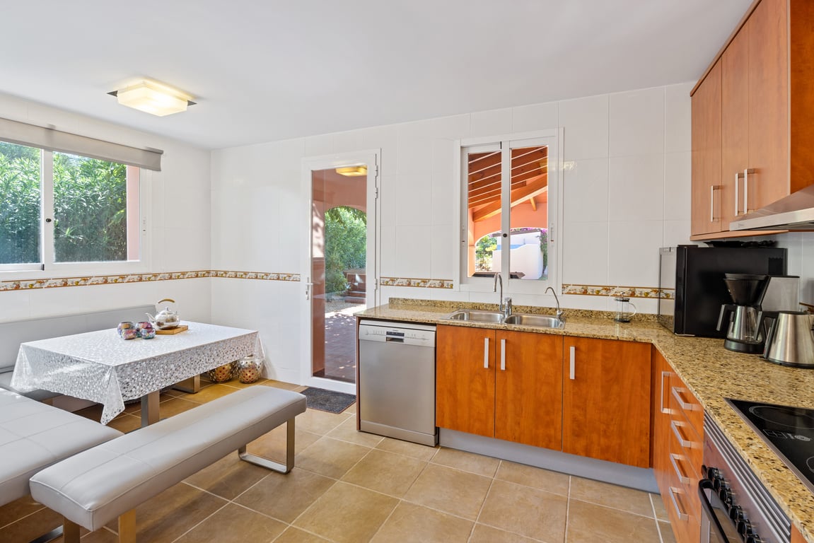 Cuisine fonctionnelle en L avec plans de travail en granit, meubles en bois et coin repas intégré. Une porte vitrée offre un accès direct vers l'extérieur.