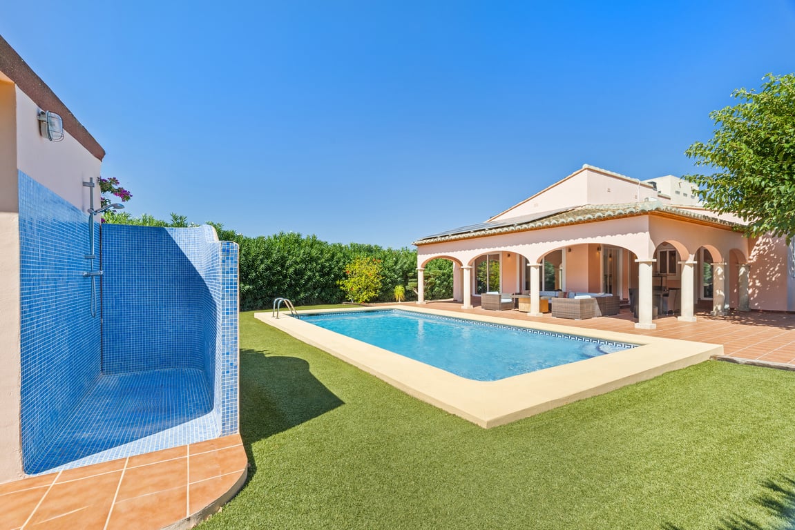 Espace extérieur avec piscine, douche en mosaïque bleue et naya à arches. Le toit est équipé de panneaux solaires, le tout entouré d'un gazon synthétique soigné.