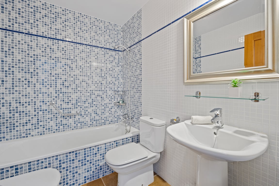 Salle de bain fonctionnelle avec baignoire carrelée de mosaïques bleues et blanches, lavabo sur colonne et grand miroir à cadre argenté.