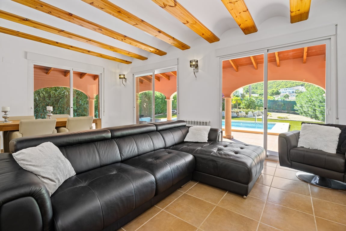 Salon lumineux avec poutres apparentes, canapé d'angle en cuir noir et grandes baies vitrées coulissantes ouvrant sur la naya et la piscine.