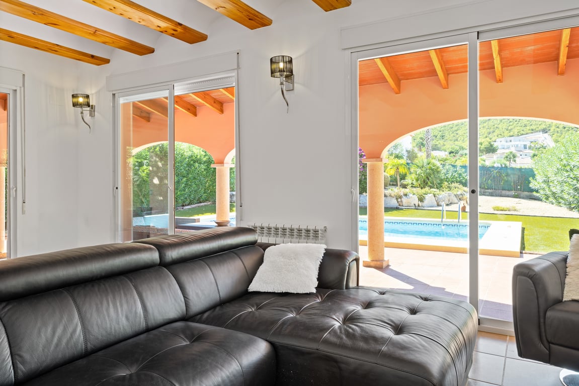 Salon lumineux avec poutres en bois apparentes, canapé en cuir noir et grandes baies vitrées offrant une vue directe sur la piscine et la naya.