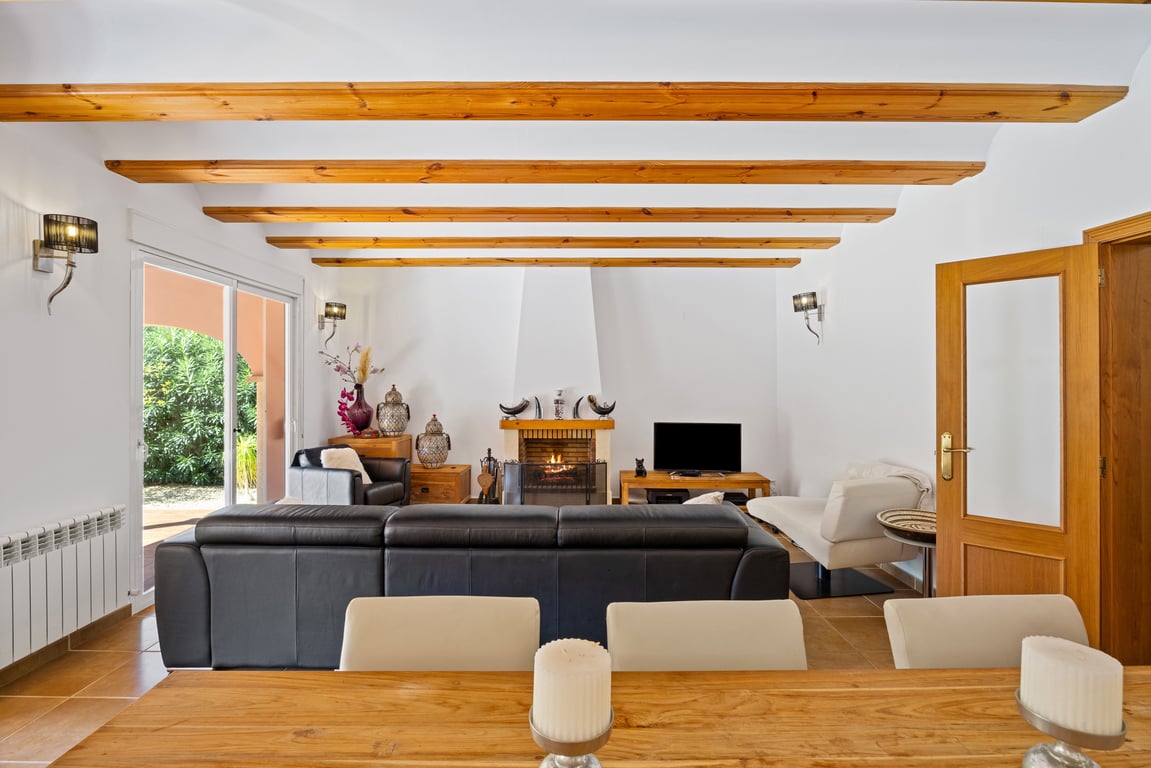 Salon lumineux avec poutres en bois apparentes, cheminée centrale et accès direct à la naya par des baies vitrées coulissantes. Sols en céramique et chauffage par radiateurs.