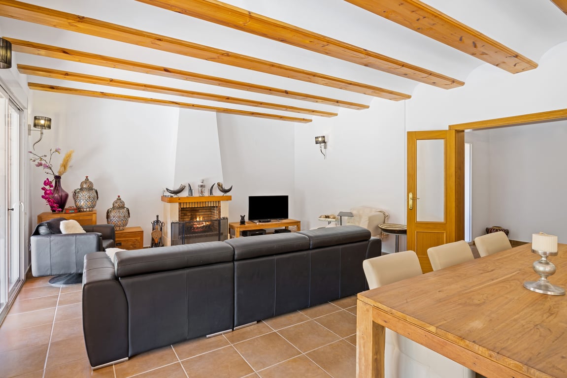 Vaste salon-salle à manger avec poutres apparentes, sol en céramique et cheminée centrale, offrant un accès direct à la terrasse extérieure.