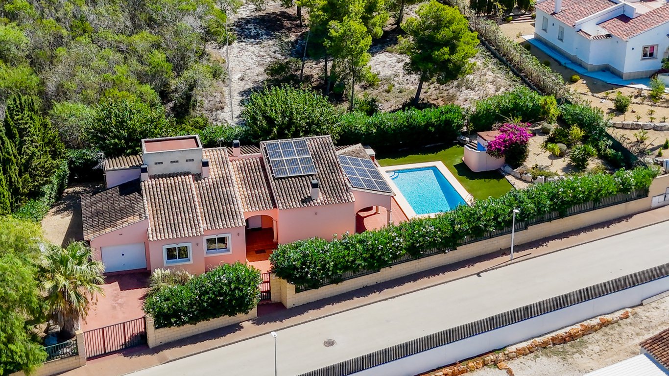 Vue aérienne de la villa montrant les panneaux solaires sur le toit, la piscine privée, le jardin avec gazon synthétique et l'accès sécurisé avec garage et portail.
