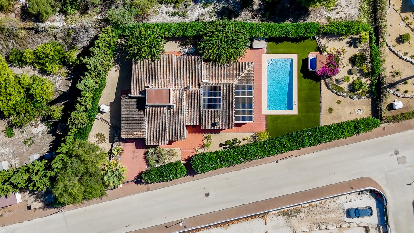 Vue aérienne de la villa montrant les panneaux solaires sur le toit, la piscine rectangulaire, le gazon synthétique et le jacuzzi entouré de haies verdoyantes.