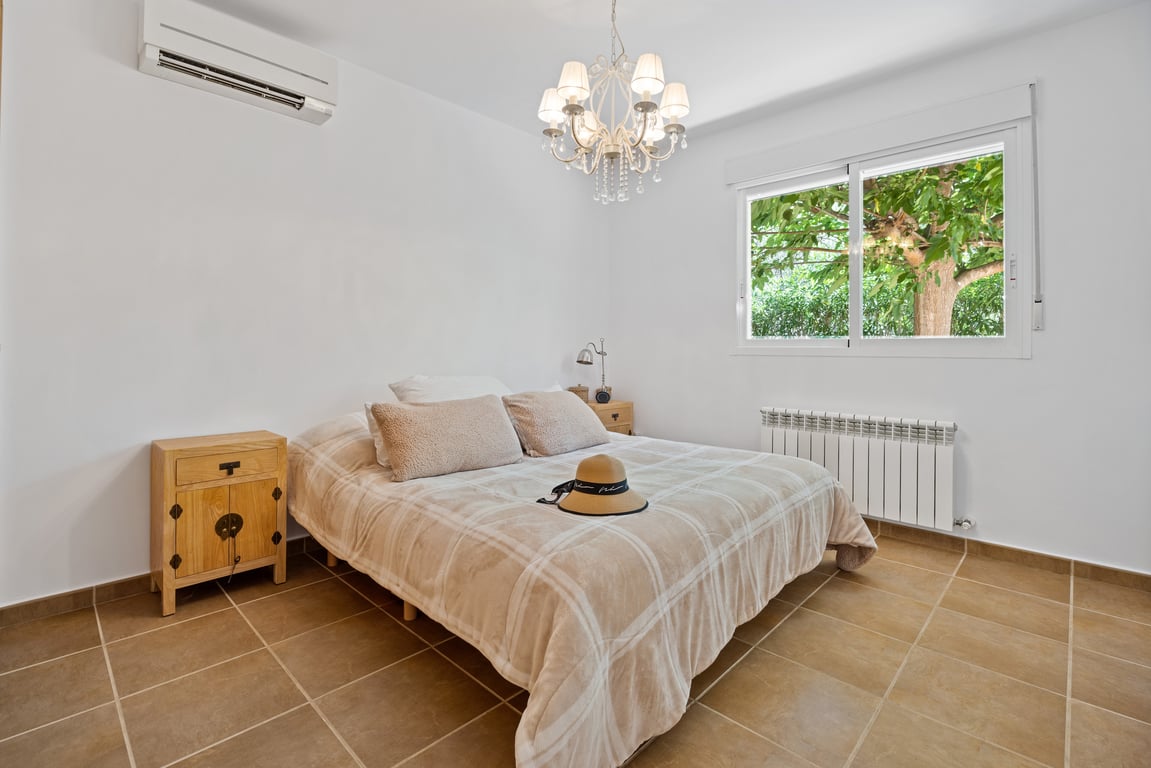 Chambre avec sol en grès, climatisation split et radiateur. Une fenêtre laisse entrer la lumière naturelle avec vue sur les arbres du jardin méditerranéen.