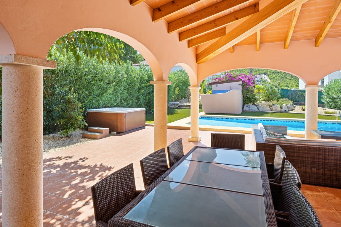 Naya couverte avec poutres en bois et colonnes en pierre, offrant une vue sur la piscine en mosaïque, le jacuzzi extérieur et le jardin paysager.