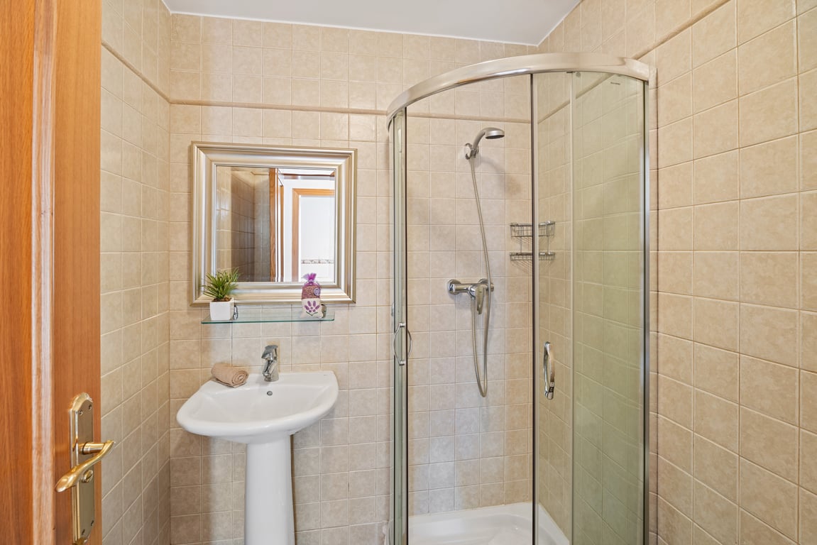 Salle de bains équipée d'une cabine de douche d'angle en verre, d'un lavabo sur colonne et de murs carrelés dans des tons beiges.