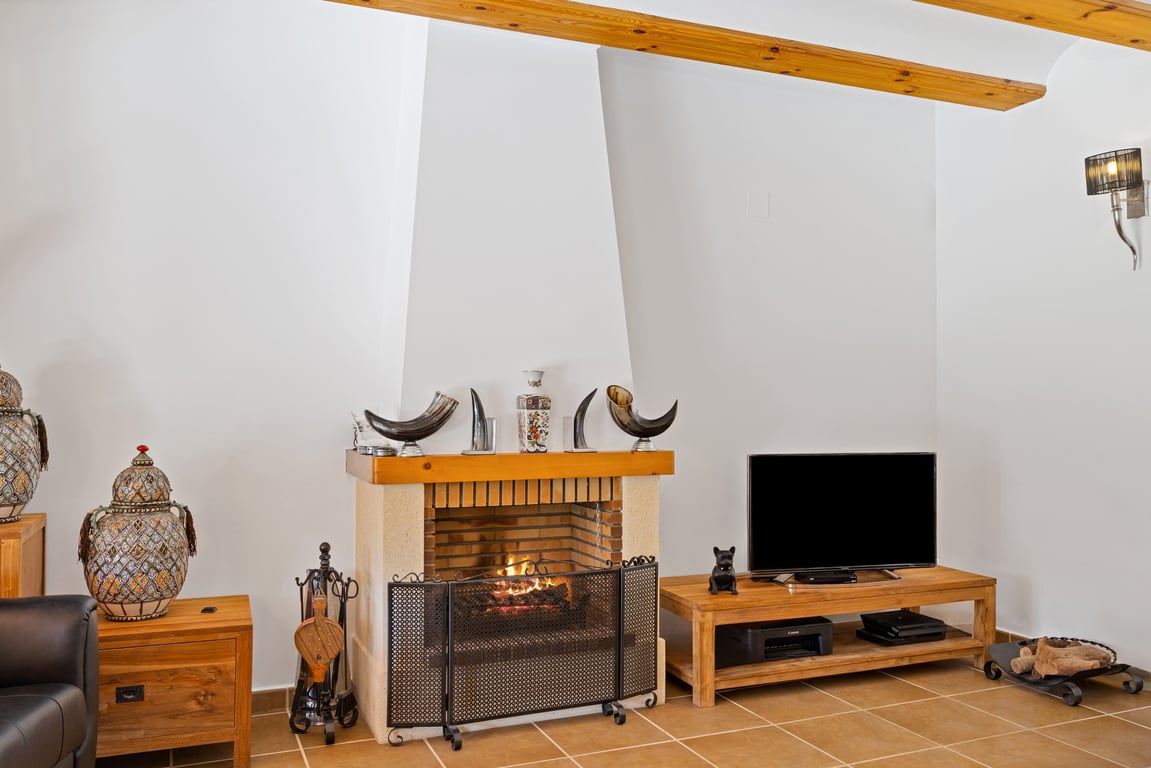 Salon avec cheminée traditionnelle, poutres en bois apparentes au plafond et sol en grès. L'espace comprend un mobilier en bois et une décoration soignée.