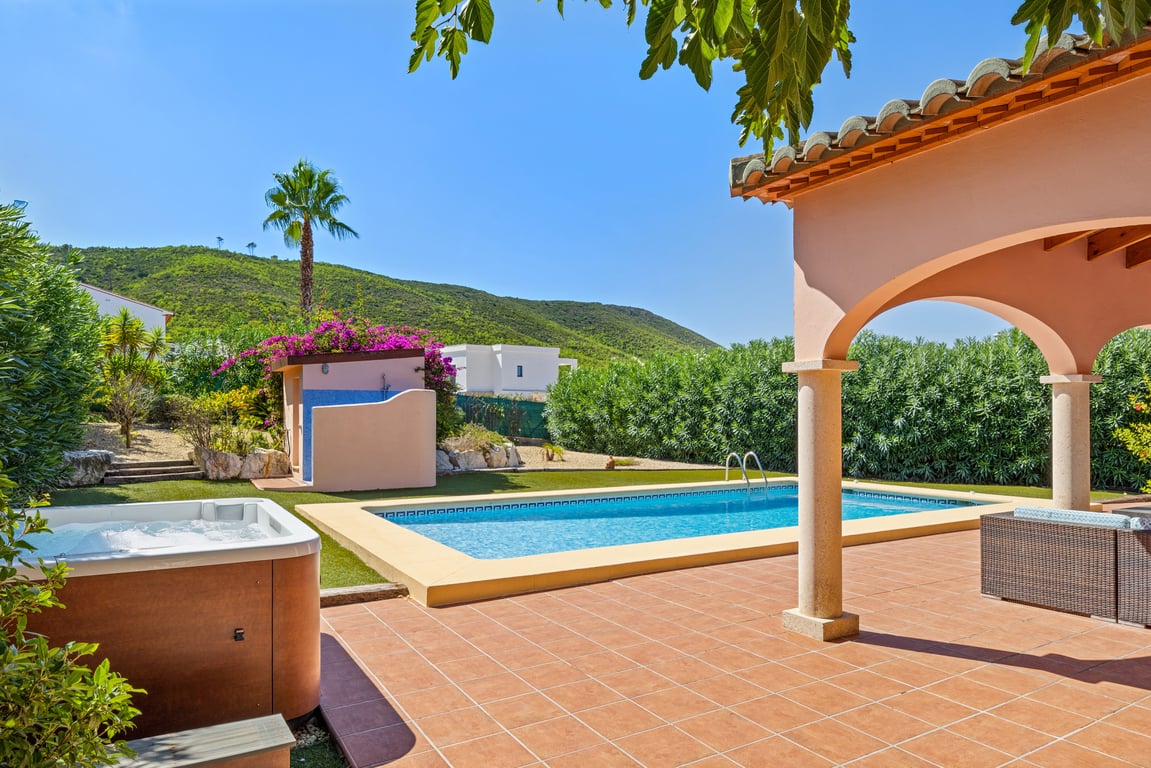 Terrasse extérieure avec piscine en mosaïque, jacuzzi indépendant et naya à arches, entourée d'un jardin avec gazon synthétique et vue sur la montagne.