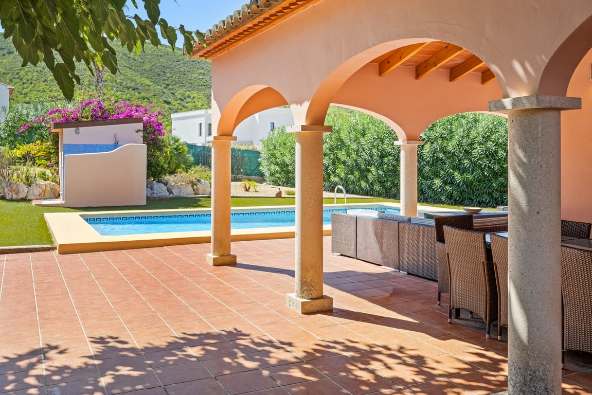 Vaste naya couverte avec arches et colonnes en pierre, donnant sur la piscine en mosaïque et un jardin avec vue sur les montagnes.