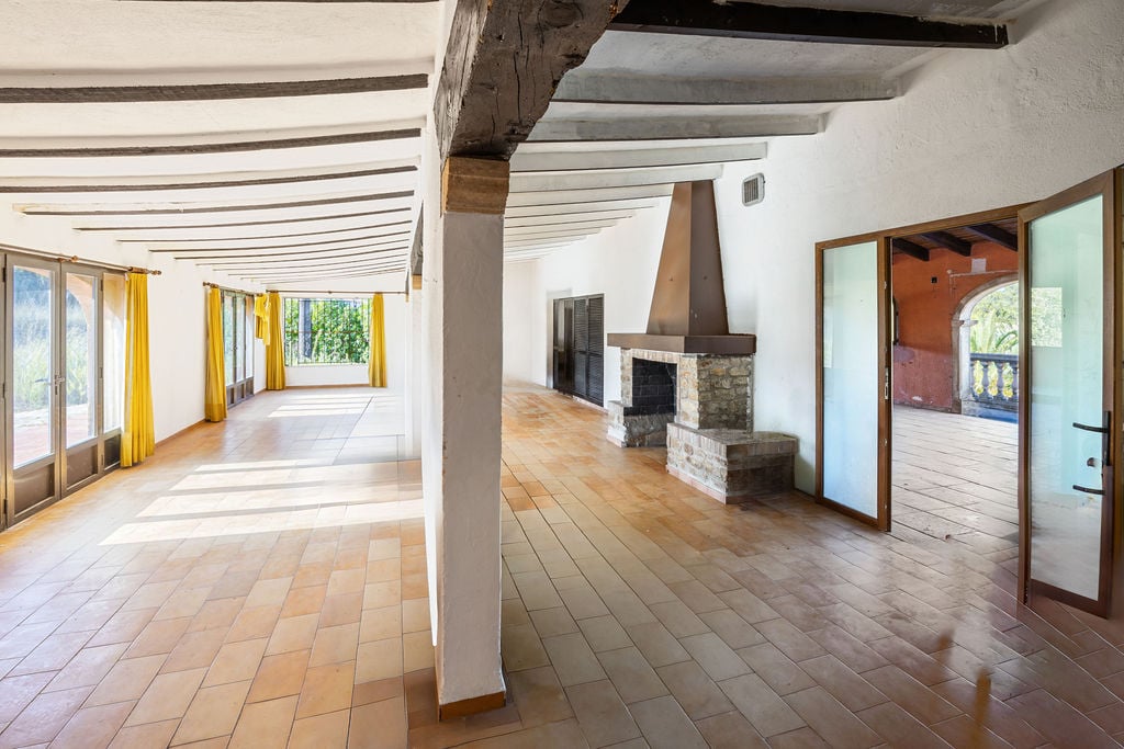 Grand salon rustique avec sol en terre cuite, poutres en bois apparentes et cheminée en pierre. Les larges ouvertures vitrées apportent une luminosité naturelle constante.