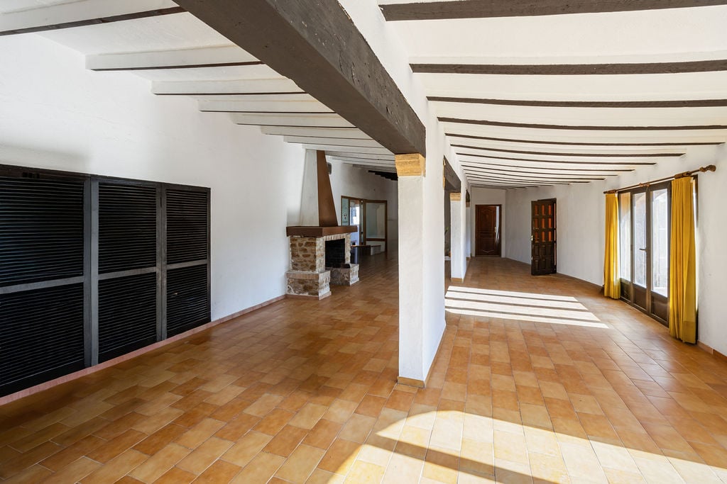 Grande pièce à vivre avec poutres apparentes en bois sombre, sol en terre cuite et cheminée en pierre. L'espace bénéficie d'une belle luminosité naturelle.