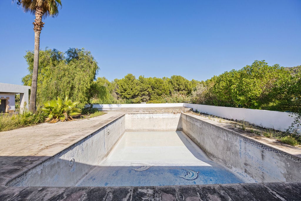 Grande piscine rectangulaire à rénover, dotée de détails en mosaïque et entourée d'une terrasse en pierre au cœur d'un jardin méditerranéen arboré.
