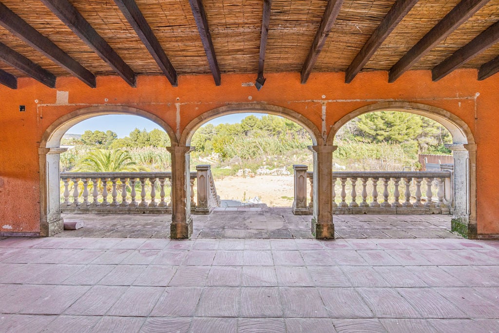 Terrasse couverte avec arches en pierre, plafond en poutres de bois et canisse, offrant une vue sur la végétation méditerranéenne à travers une balustrade classique.