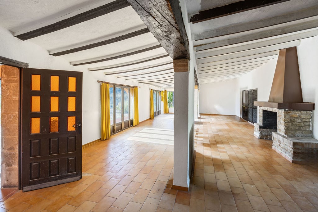 Vaste salle rustique avec poutres apparentes en bois sombre, sol en terre cuite et une cheminée en pierre naturelle avec hotte métallique.
