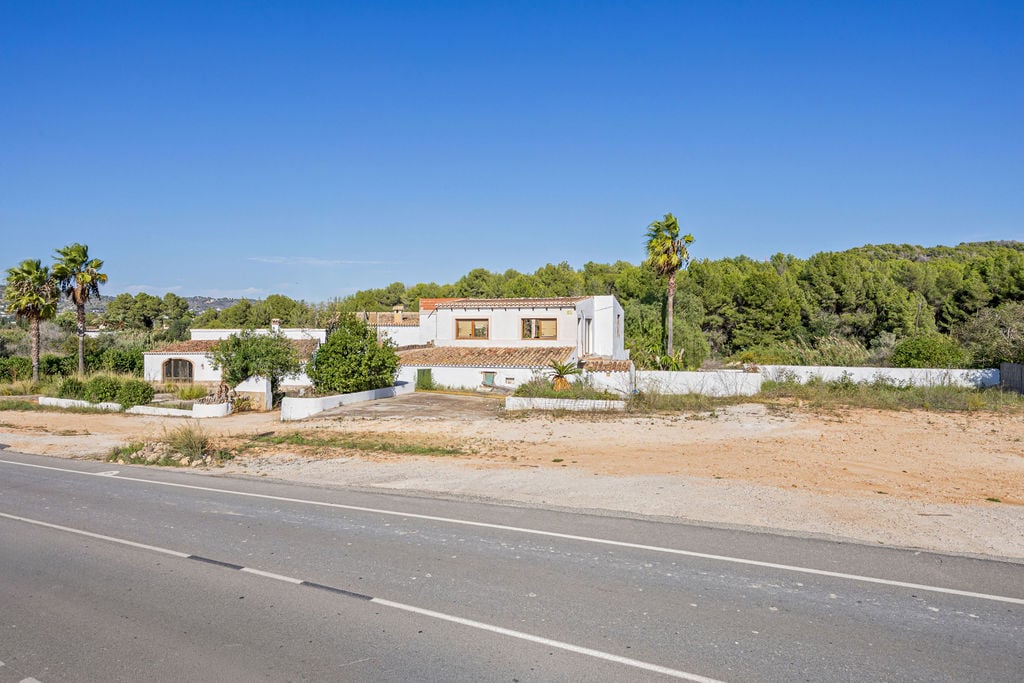 Vue extérieure de la finca traditionnelle avec façades blanches et toits en tuiles, bordée par une route d'accès et entourée d'une forêt de pins méditerranéens.