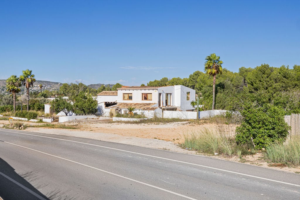Vue extérieure de la propriété depuis la route, mettant en valeur l'architecture traditionnelle aux murs blancs et toits de tuiles, entourée de pins et palmiers.