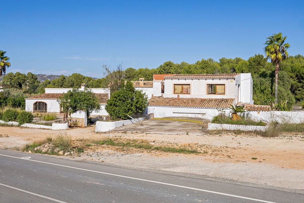 Vue extérieure d'une bâtisse méditerranéenne traditionnelle avec murs blancs et toits en tuiles, située sur un terrain spacieux en bordure de route.