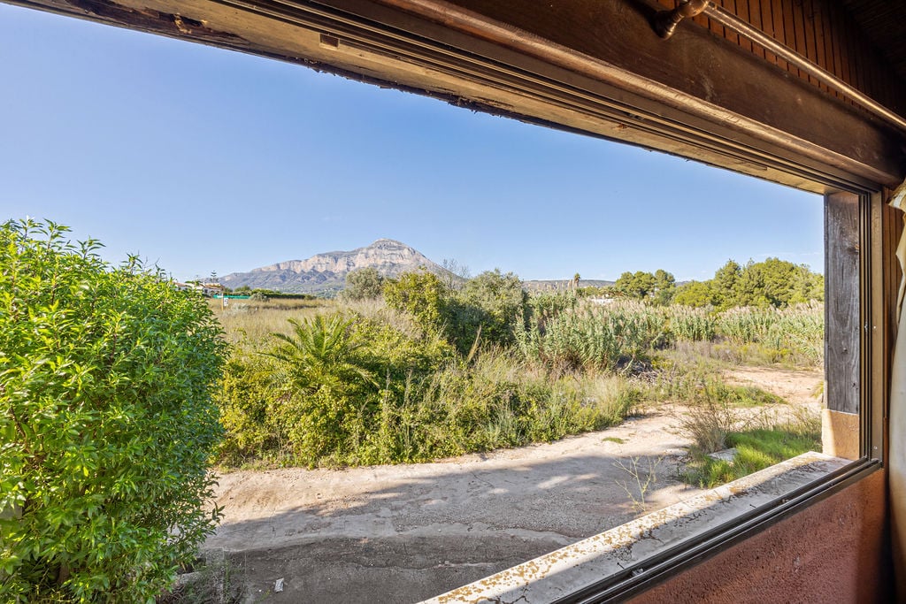 Vue panoramique sur le mont Montgó depuis une fenêtre à cadre en bois. L'extérieur se compose d'une végétation méditerranéenne dense sous un ciel dégagé.