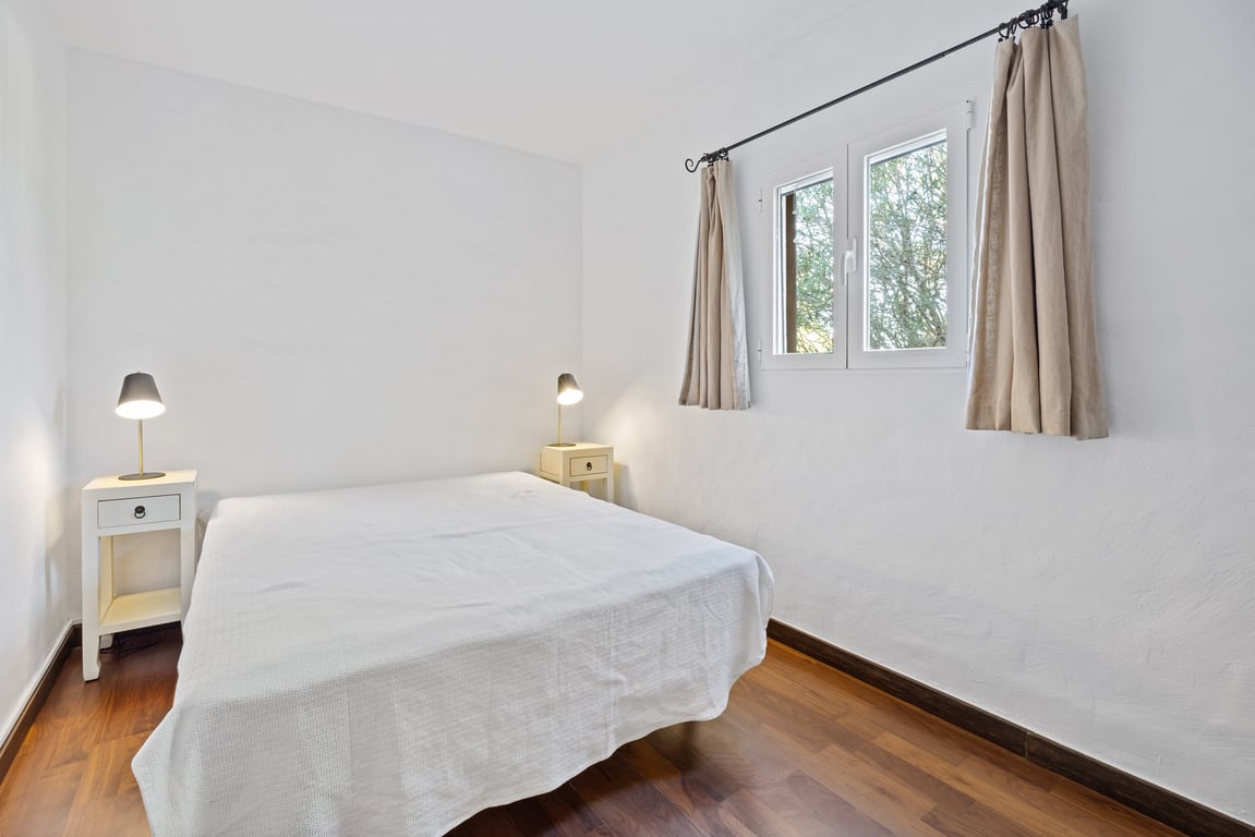 Chambre lumineuse avec parquet, murs blancs et fenêtre à double vitrage offrant une vue sur la verdure. Équipée de tables de chevet et de lampes de lecture.