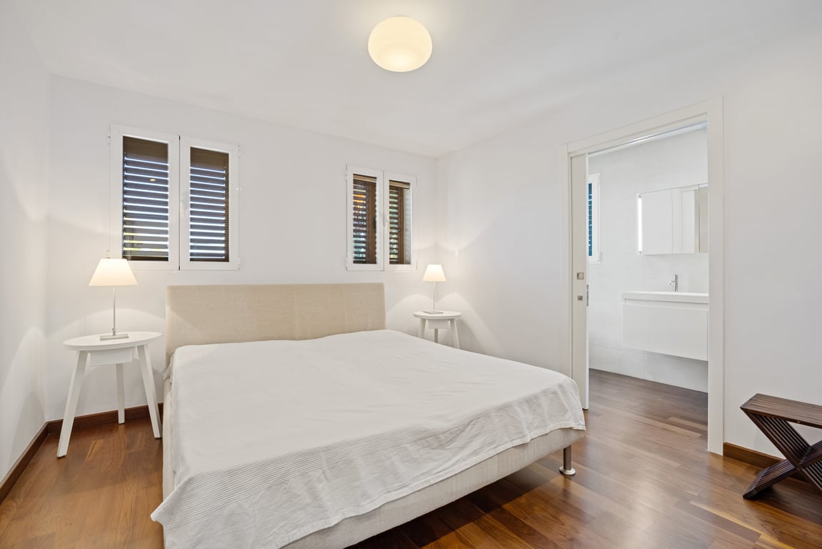 Chambre rénovée avec parquet foncé, tête de lit en tissu et porte coulissante ouvrant sur une salle de bains privative moderne et lumineuse.