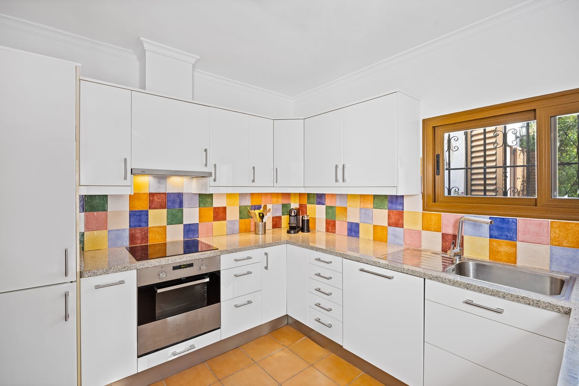 Cuisine équipée avec placards blancs, plan de travail en granit et crédence en carreaux de céramique artisanaux colorés. Comprend un four encastré et une fenêtre en bois.