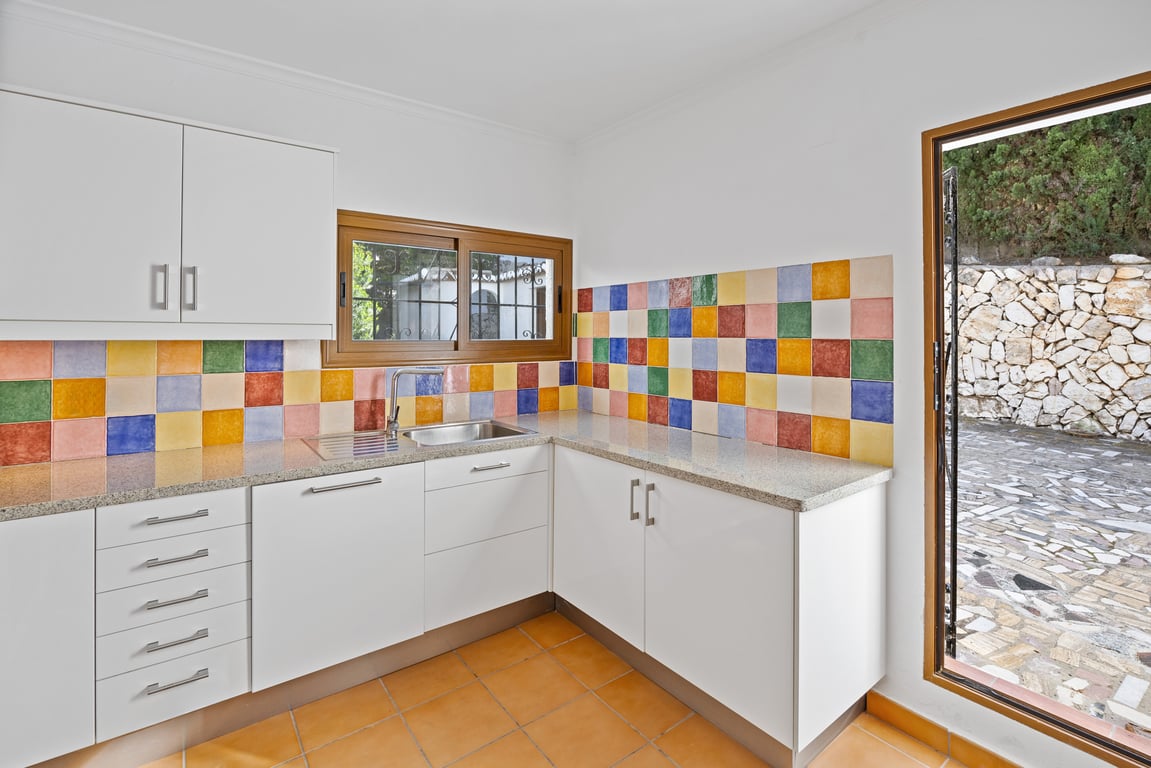 Cuisine équipée de meubles blancs, d'un plan de travail en granit et d'une crédence colorée. Elle dispose d'un sol en terre cuite et d'un accès direct vers la terrasse.