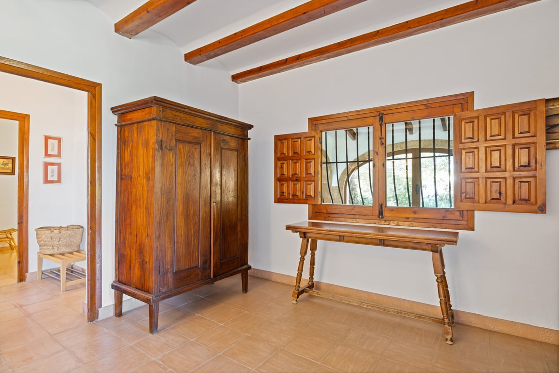 Entrée avec poutres apparentes, sol en céramique artisanale et mobilier en bois massif. La fenêtre dispose de volets traditionnels et de grilles de sécurité.