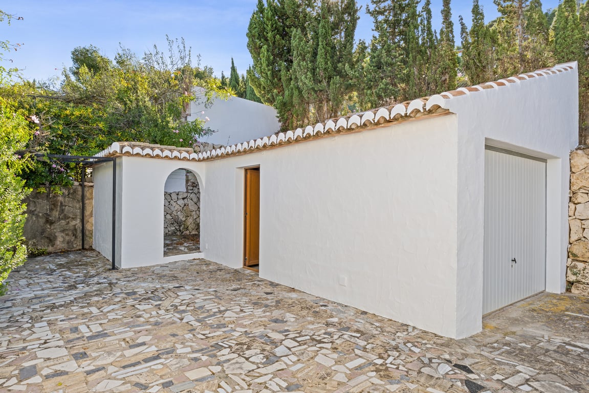 Garage indépendant et espace de service avec façade blanche, toiture en tuiles canal et sol en mosaïque de pierre, entourés de cyprès matures.