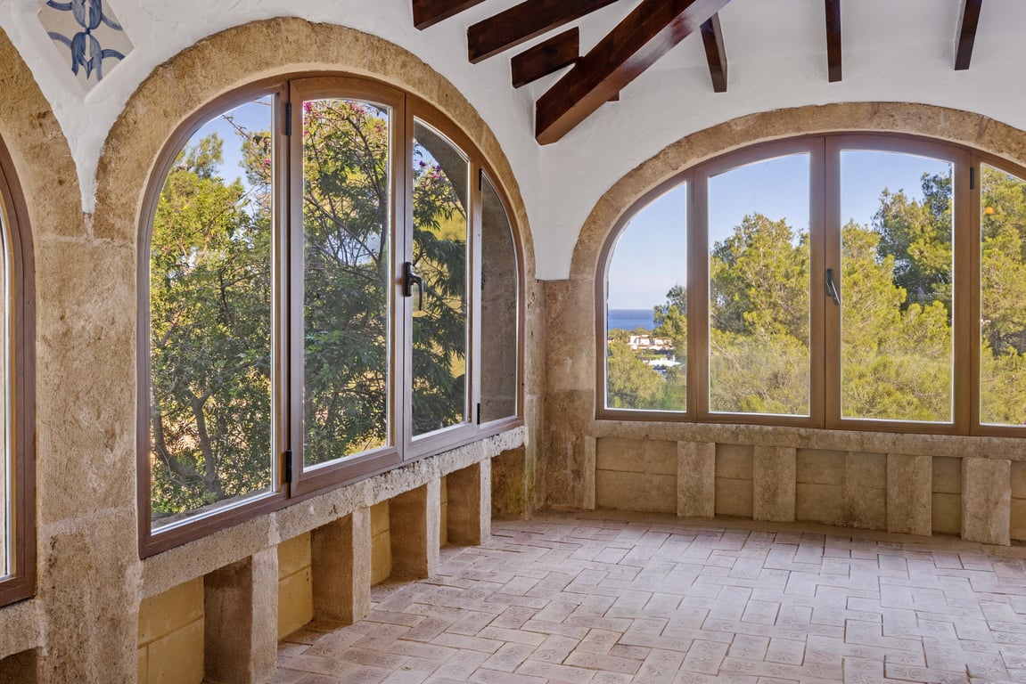 Naya vitrée avec arches en pierre Tosca, poutres en bois apparentes et sol en terre cuite. Les fenêtres offrent une vue sur la mer et la végétation environnante.