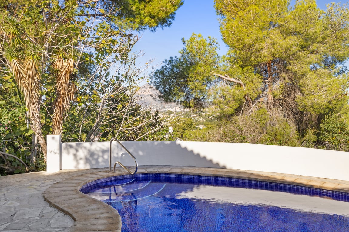 Piscine en mosaïque bleue avec rampe en métal, entourée d'une terrasse en pierre naturelle et d'une végétation méditerranéenne offrant une vue sur le Montgó.