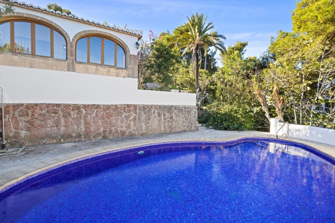 Piscine privée avec mosaïque bleue, bordée d'un mur en pierre naturelle et d'une terrasse, avec la naya aux fenêtres arquées entourée de végétation.