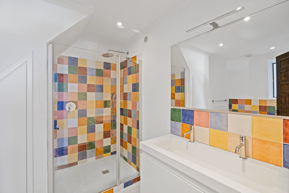 Salle de bain moderne avec double vasque et douche vitrée, caractérisée par ses carreaux de céramique multicolores et son éclairage au plafond encastré.