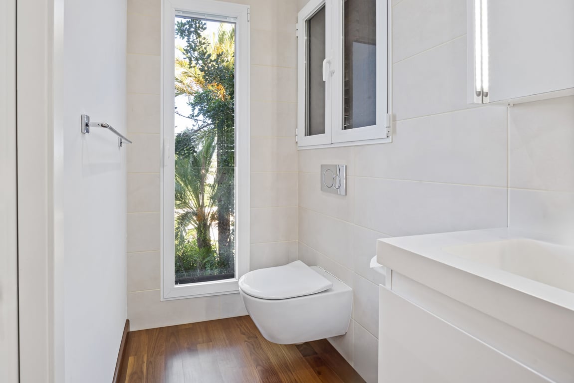 Salle de bain rénovée avec WC suspendu, sol en bois et fenêtre verticale offrant une vue sur la végétation extérieure.