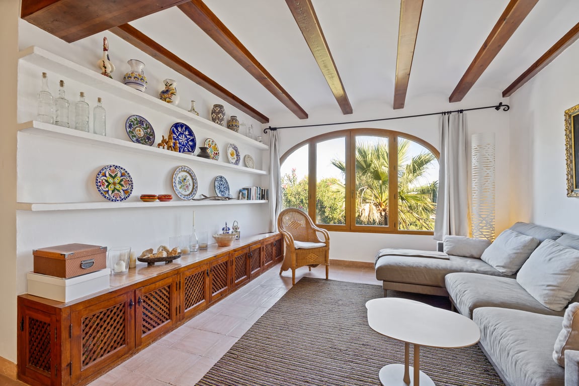Salon avec poutres en bois apparentes, étagères intégrées ornées de céramiques et grande fenêtre cintrée offrant une vue sur le jardin.