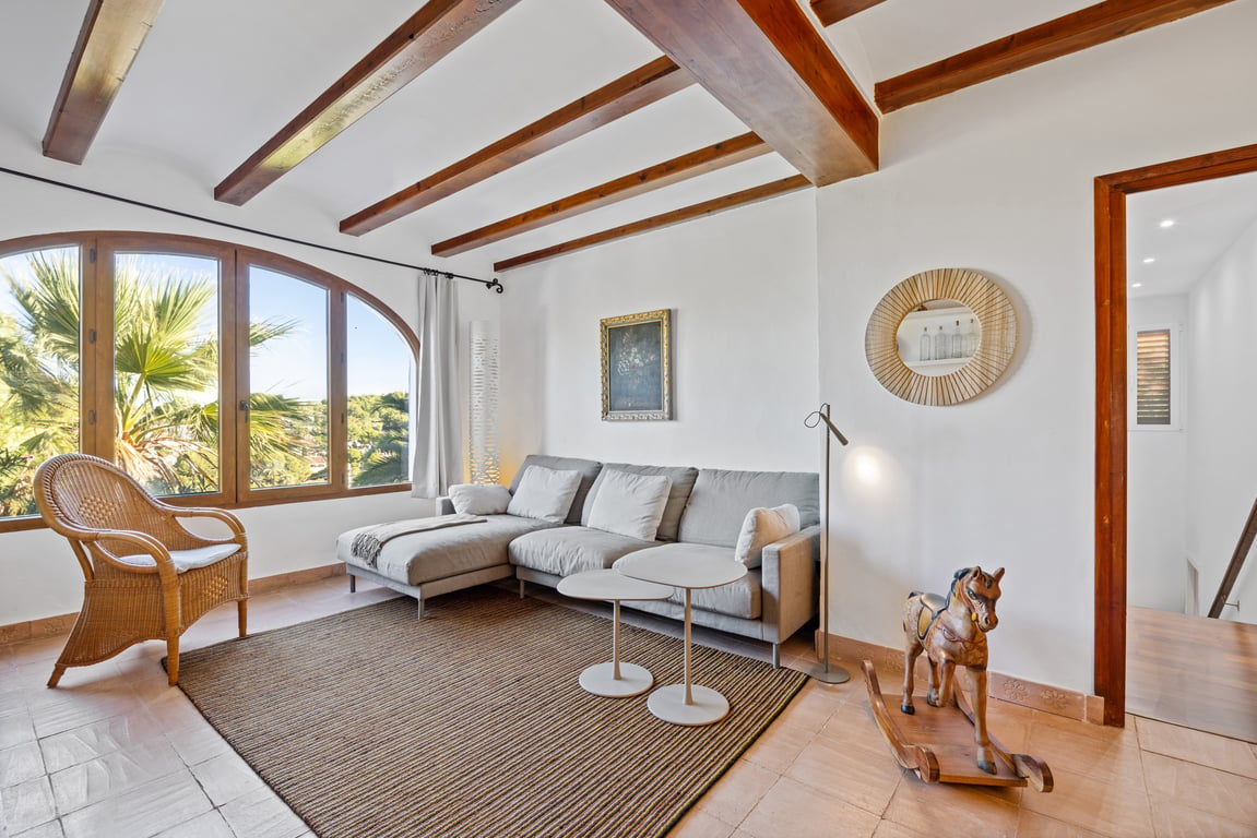 Salon lumineux avec poutres en bois apparentes, sol en terre cuite et une grande fenêtre en arc offrant une vue sur la végétation.