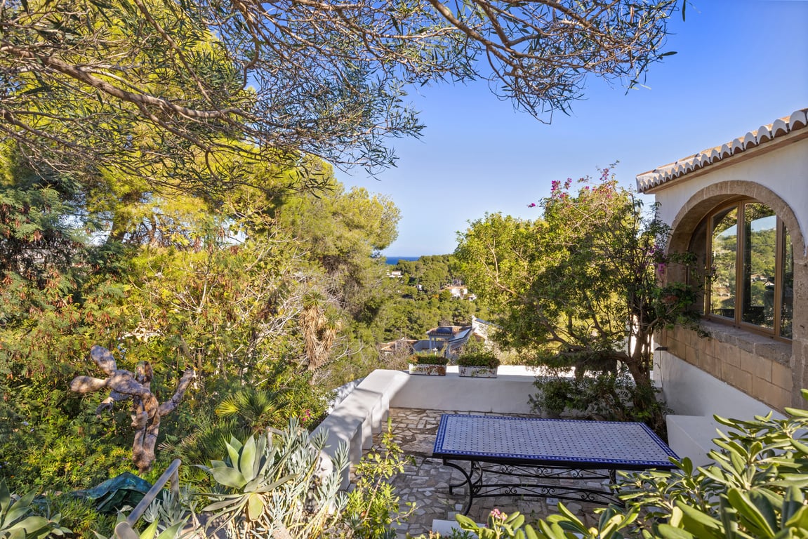 Terrasse extérieure avec pavage en pierre naturelle et table en mosaïque, entourée de végétation méditerranéenne offrant une vue sur la mer à travers les pins.