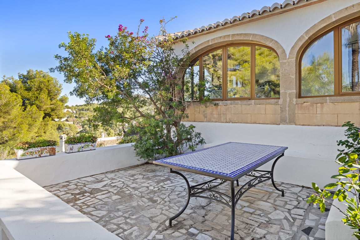 Terrasse extérieure avec revêtement en pierre naturelle, table en céramique artisanale et arches en pierre de Tosca encadrant la naya vitrée ensoleillée.