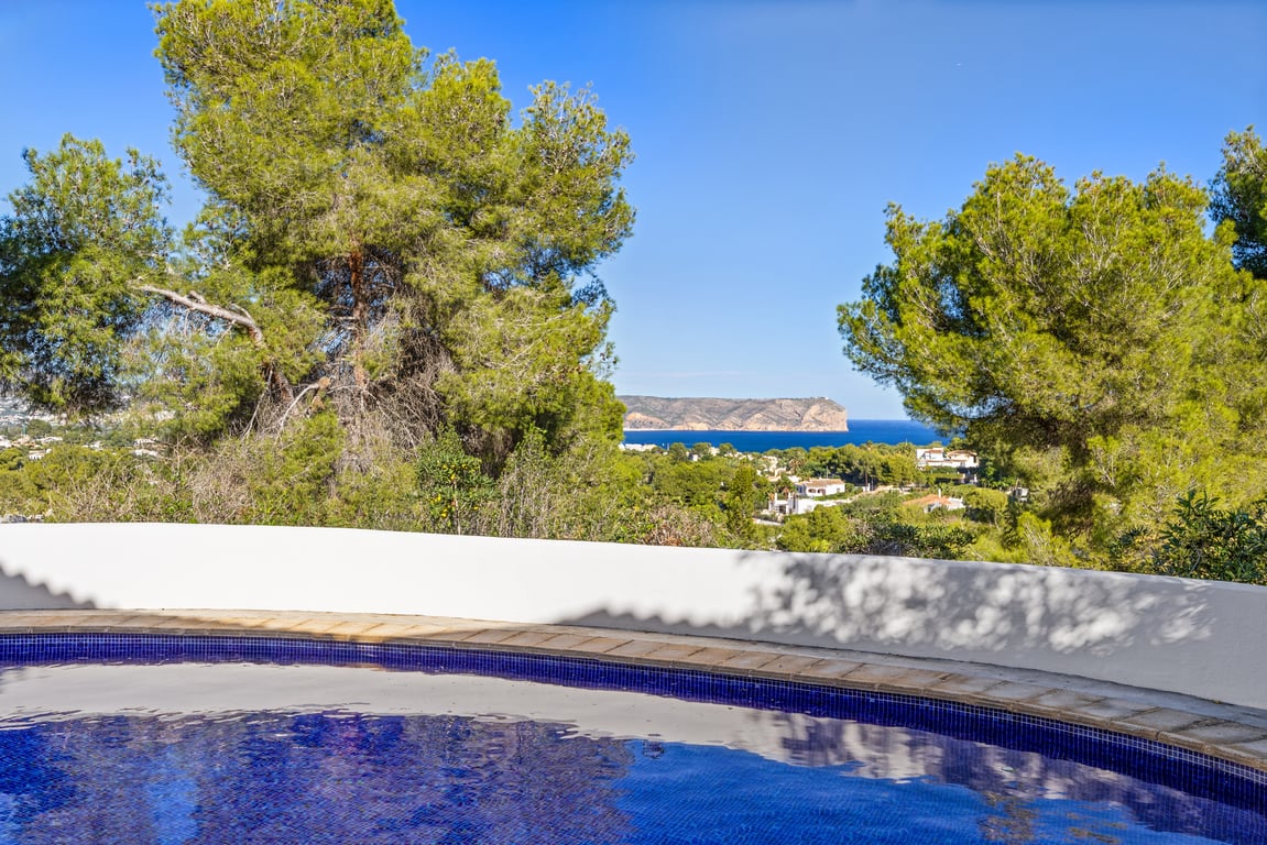 Vue depuis la piscine privée sur la mer Méditerranée et les falaises, entourée de pins matures et d'un muret blanc incurvé.