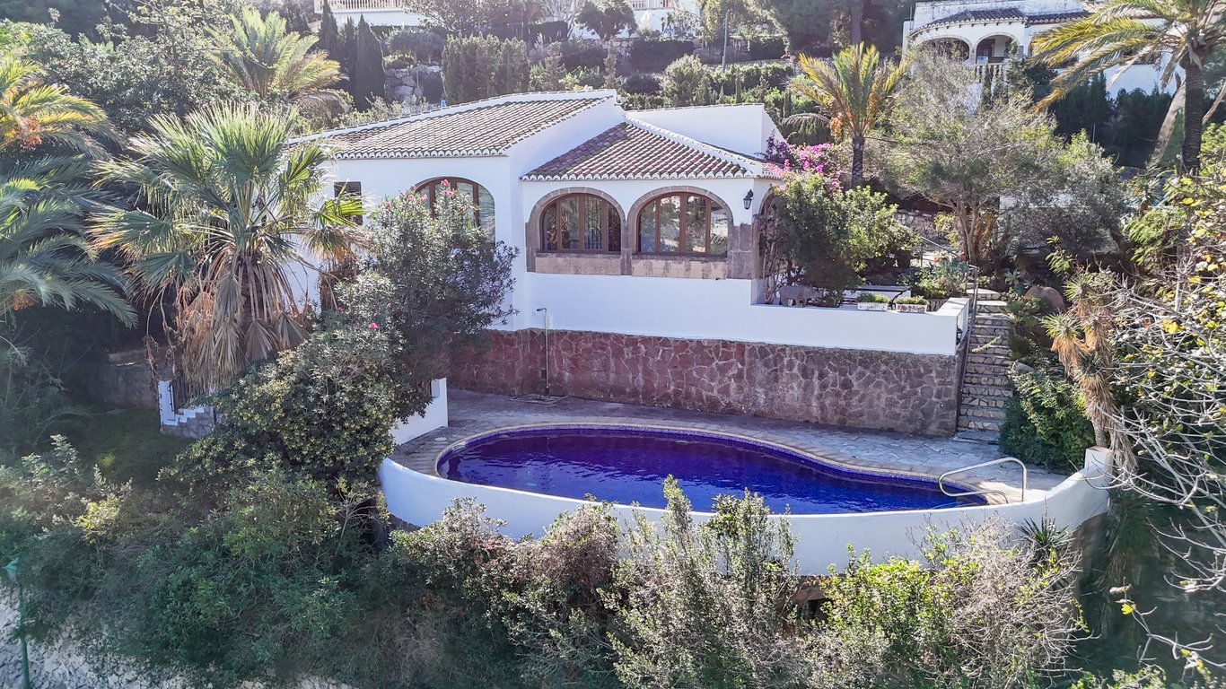 Vue extérieure de la villa avec piscine privée ovale, murs en pierre naturelle et naya traditionnelle aux arches en pierre de Tosca.