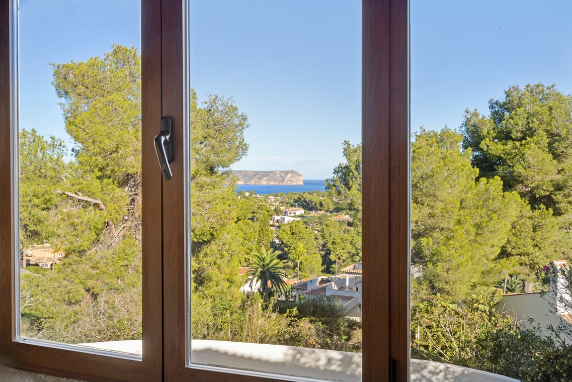 Vue panoramique sur la mer et le cap à travers une fenêtre en bois à double vitrage, surplombant une pinède méditerranéenne.