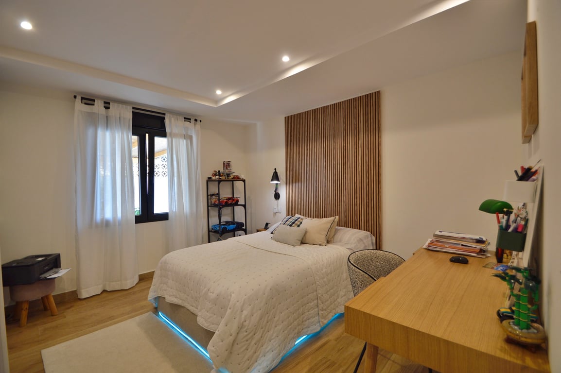 Chambre contemporaine avec tête de lit en lattes de bois, bureau intégré et lit avec éclairage LED bleu. Comprend un sol en bois et des spots de plafond encastrés.