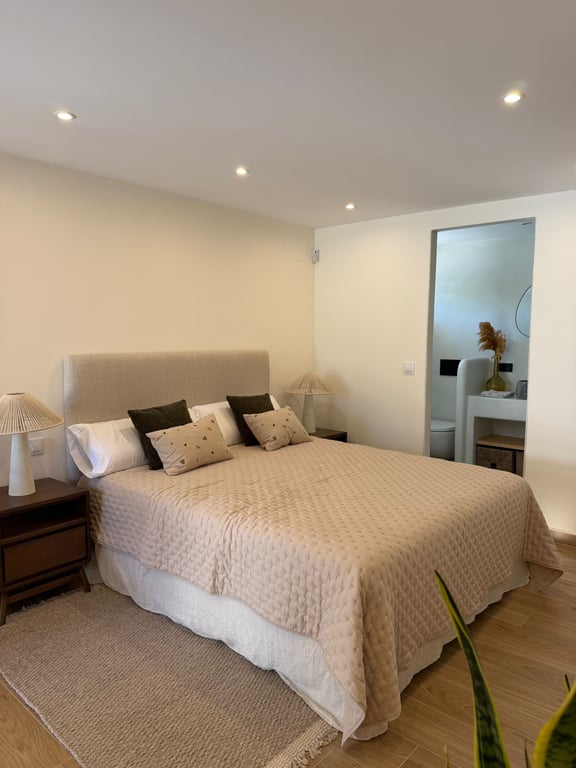 Chambre lumineuse avec lit double, tête de lit rembourrée et tables de chevet en bois. Salle de bain attenante visible avec finitions modernes et éclairage encastré.